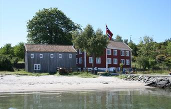 bilde av Merdøgaard og en grå låve. foran bygningene ligger en fin sandstrand.