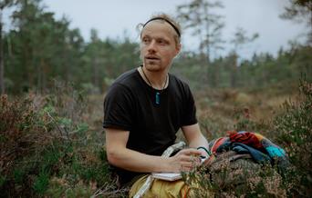 artisten Moddi sittende i lyngen med penn og papir.