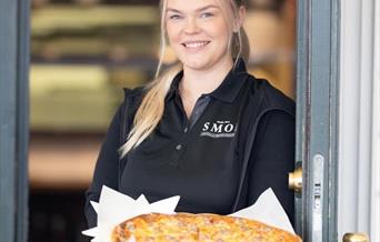 Blid servitør fra SMOI som står i døra og holder en deilig pizza