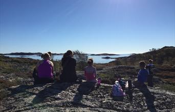 Bilde av seks mennesker som sitter på stein og ser utover mot havet, sola skinner ned på dem, en dame tar bilde av utsikten