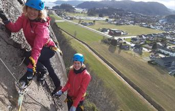 To personer, iført sikkerhetsutstyr, klatrer Via Ferrata i Lyngdal. I bakgrunnen kan man se boligområdet, grønt landskap, og fjell.