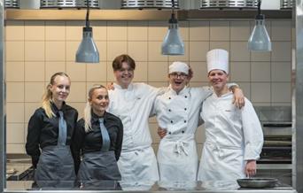 Tre unge kokkeelever i hvite kokkeklær og to unge servitriser i svarte arbeidsklær fotografert på skolekjøkkenet på Sam Eyde vgs.