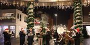 Et korps med barn og unge i nisseluer spiller julemusikk under julebyen Lyngdal i stemningsfull kveldsbelysning.