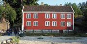 fronten på Merdøgaard skjærgårdsmuseum. et rødt skipperhus fra 1736 i to etasjer. Foran huset er det sandstrand og fergebrygge.
