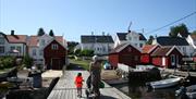 brygga hvor fergene legger til på Merdø. på brygga ses en mor med barnevogn og et barn med oransje flytevest. i bakgrunnen ses bebyggelsen med gamle hvite trehus og røde sjøboder.