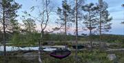 en rosa hengekøye hengende mellom 2 trær midt på bildet, og en svart litt lengre bak, en dame sitter ved vannet