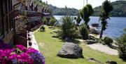 feriehus med rosalilla blomster i blomsterkasse på alle terrassene på venstre side med nyklipt hage og vann på høyre side. Norsk vimpel på flaggstang. skyfri himmel.