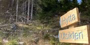 Treskilt med blå skrift "Hytta" og "Utsikten" pekende oppover en sti i skogen.