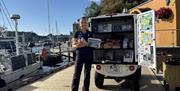 En smilende havnevert, som står foran turistinformasjon bilen i gjestehavna, med rundstykker til båtgjestene i hånda