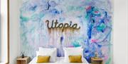hotellvegg med streetart på veggen over sengegavlen i turkis, blått og litt rosa med ordet Utopia i gullskrift midt på det hele.