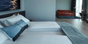 Hotellrom med dobbeltseng, andre siden av rommet er det en rød sofa med lite bord oppå svart teppe, en pult med stol. Stort vindu med mørkegrå gardiner.