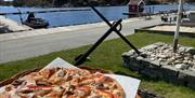 Deilig pizza med kjøtt, løk, paprika på et fat med brygga og sjøen i bakgrunnen.