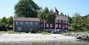 bilde av Merdøgaard og en grå låve. foran bygningene ligger en fin sandstrand.