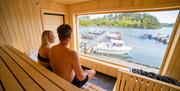 Mann og dame som sitter i saunaen på Tregde, med utsikt ut mot havet og båtene som ligger langs brygga