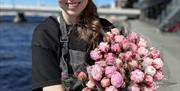 Smilende ansatt fra blomsterbutikken Floriss, viser frem flotte blomster, med Mandals elva i bakgrunnen