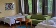 Firemanns rom, der det er en grønn sofa i midten med en dobbeltsenger på hver sin side. Gule vegger, og vinduer langs hele veggen med mørkegrå gardiner.
