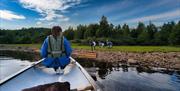 Kvinne i kano på vannet og tre geiter på land