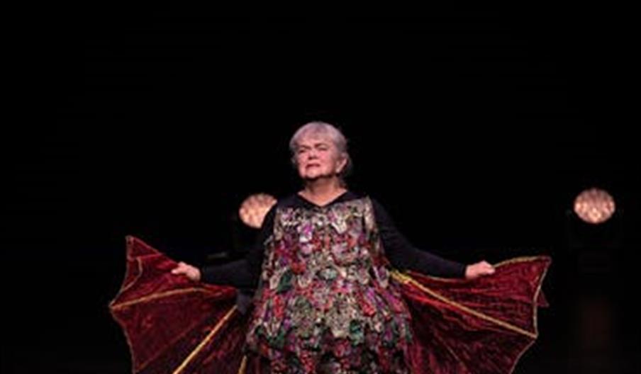 Anne Marit Jacobsen på scenen i forestillingen «Alle fugler», iført fargerik drakt og med scenelys i bakgrunnen. Anne Marit Jacobsen på scenen i forestillingen «Alle fugler», iført fargerik drakt og med scenelys i bakgrunnen.